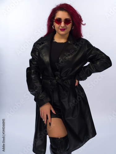 Stylish young girl with brown hair on a white background. She is dressed in a black leather raincoat and a skirt above the knee. Wears sunglasses, piercings.