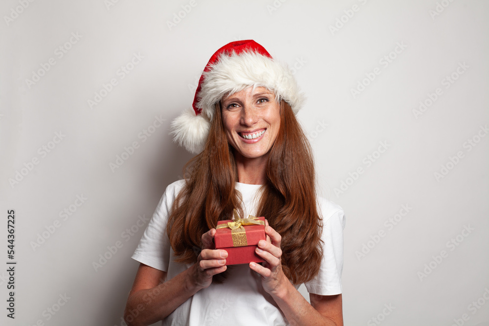 Portrait of cheerful smiling mature woman in Christmas hat holding red gift box with gold ribbons against white studio wall background Portrait of cheerful smiling mature woman in Christmas hat holding red gift box with gold ribbons against white studio wall background