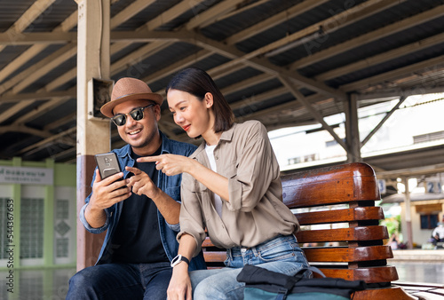 Принти на полотні Happy asian handsome man and beautiful woman backpacker wearing sunglasses, hat and backpack, They sitting together and exiting when received some message on cell phone in the train station