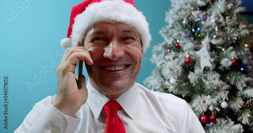 Closeup smiling man in Christmas Hat Santa Clausa talking mobile phone on the background of the Christmas tree.Portrait of happy business man calling phone indoor. photo