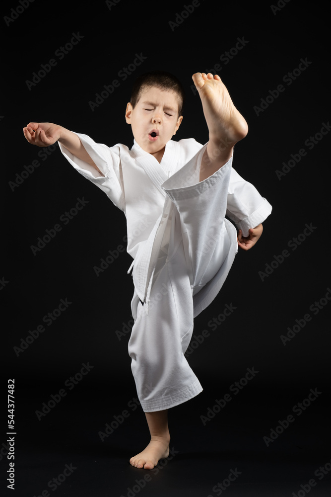 A little boy in a kimono practices karate on a black background, kicking forward. A little boy in a kimono practices karate on a black background, kicking forward.