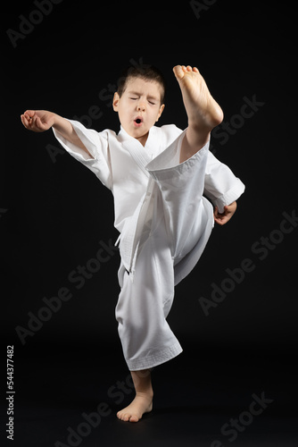 A little boy in a kimono practices karate on a black background, kicking forward.