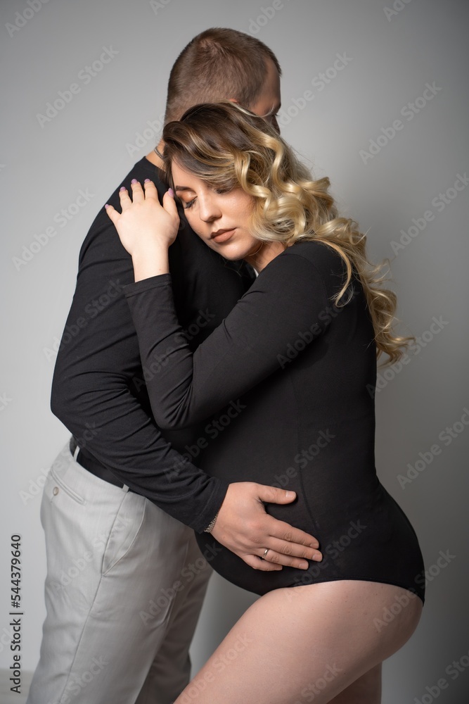 Fototapeta premium pregnant woman in a black bodysuit and a man on a gray background. Long curly hair, beautiful couple, husband and wife, future parents. stylish pregnancy. portrait