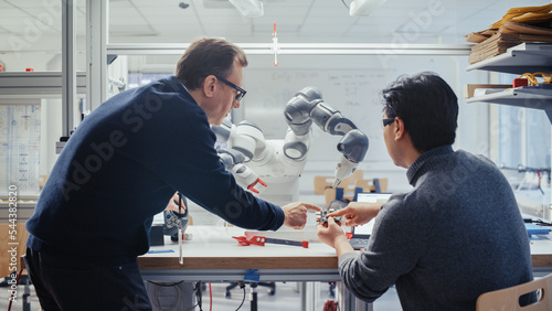 Adult Caucasian Engineer Changes Position of Robotic Arm with Controller and Having a Discussion with Young Asian Developer. Startup Lab and Innovative Technologies in Robotics Concept. photo