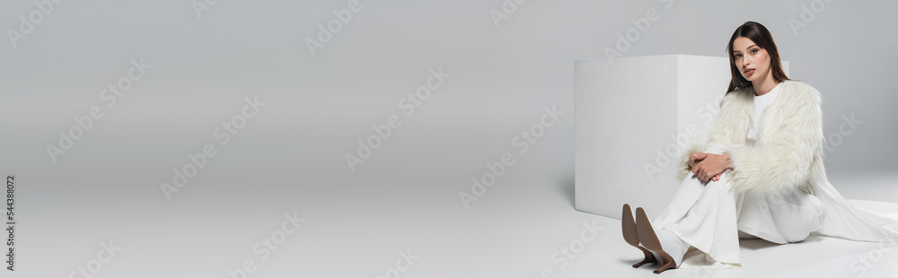 full length of stylish young woman in white faux fur jacket looking at camera near cube on grey, banner. full length of stylish young woman in white faux fur jacket looking at camera near cube on grey, banner.