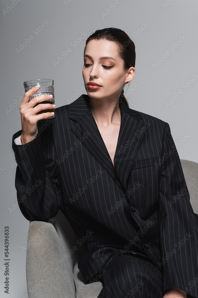 Stylish woman in striped jacket looking at glass of whiskey on armchair isolated on grey. Stylish woman in striped jacket looking at glass of whiskey on armchair isolated on grey.