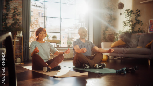 Senior Couple Meditating Together After Morning Exercises at Home in Sunny Living Room. Middle Aged Man and Woman Rest After Workout. Healthy Lifestyle, Fitness, Recreation, Wellbeing and Retirement.