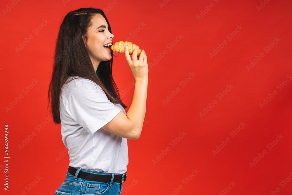 Portrait of young beautiful hungry woman eating croissant. Isolated portrait of woman with fast food over red background. Diet breakfast concept. Portrait of young beautiful hungry woman eating croissant. Isolated portrait of woman with fast food over red background. Diet breakfast concept.
