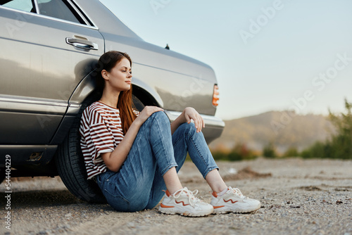 A young woman sits on the ground near her car on the side of the road and looks at the sunset © SHOTPRIME STUDIO