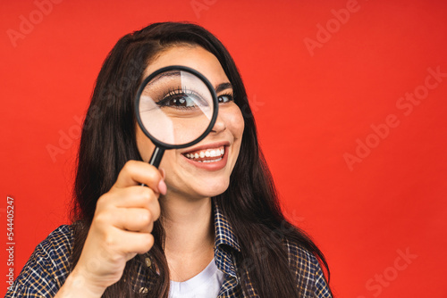 Close up portrait of amazing beautiful woman looking at camera through magnifying glass, isolated on red background. Having fun.