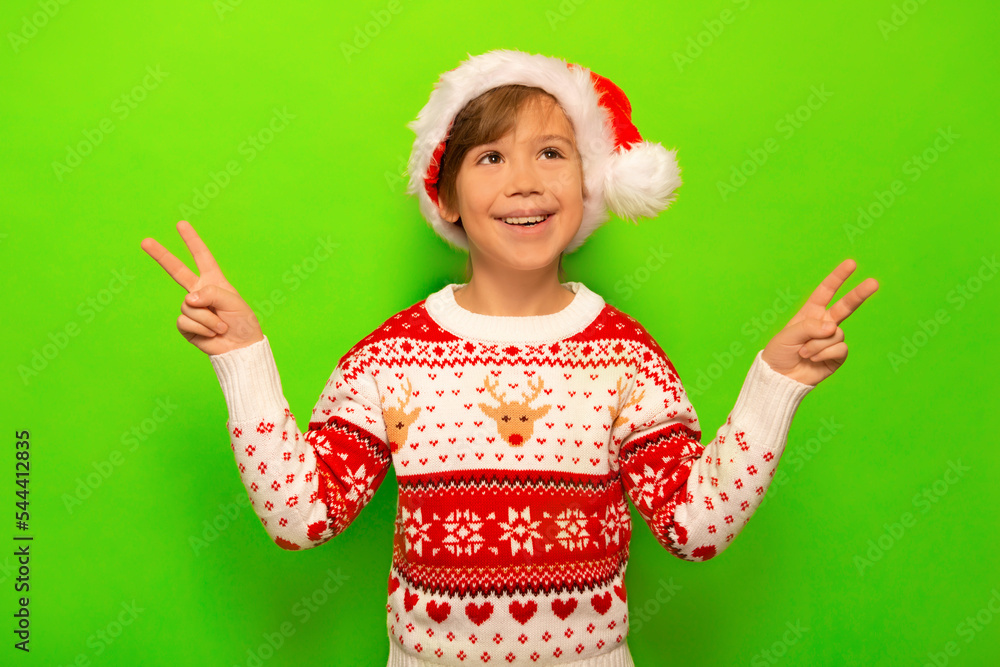 Happy smiling little boy in Santa Claus hat, Christmas sweater with deer and snowflakes pattern showing peace gesture or v sign with two fingers and looking up on a green background. Happy smiling little boy in Santa Claus hat, Christmas sweater with deer and snowflakes pattern showing peace gesture or v sign with two fingers and looking up on a green background.