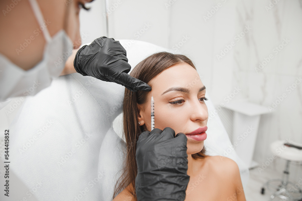 Fototapeta premium A cosmetologist makes anti-aging injections against wrinkles on the forehead on the face of a beautiful woman. Women's aesthetic cosmetology in a beauty salon.