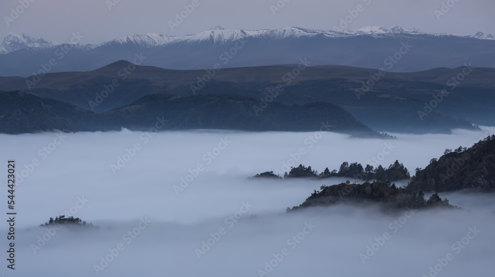Fototapeta premium Low clouds in the mountains filled the entire valley and rocky peaks and hills with trees stick out above the clouds, mountains with snow in the background, fog between the mountains on an autumn