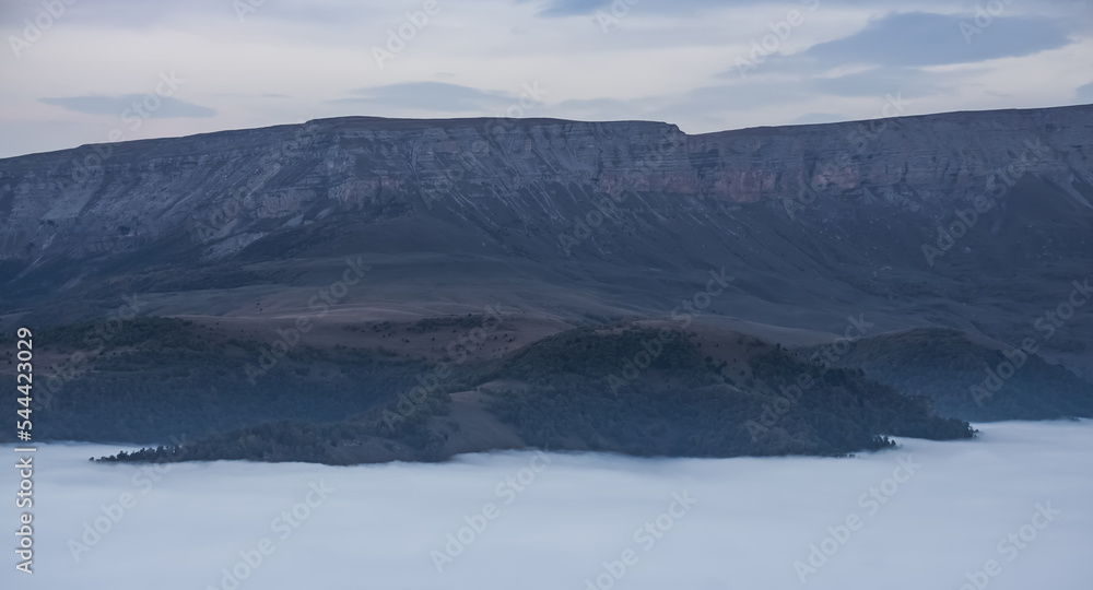 Mountain evening landscape with low clouds in the valley and a steep cliff in the background, panorama of the evening mountains