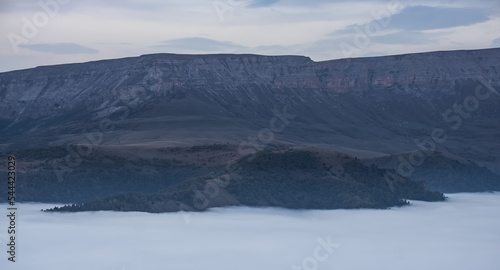 Mountain evening landscape with low clouds in the valley and a steep cliff in the background, panorama of the evening mountains