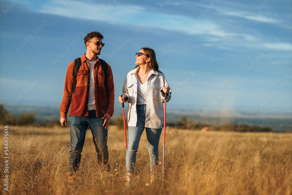A couple is chatting and hiking in big golden yellow meadow on a sunny autumn day. They wear sunglasses to protect them from sun. The girl is holding trekking poles to help her while hiking.