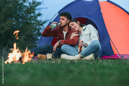 A happy couple is sitting and drinking a beverage in front of a blue tent by the camp fire. They enjoy camping in nature.