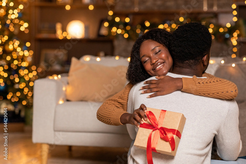 Smiling millennial african american woman hug man with present box in cozy living room interior