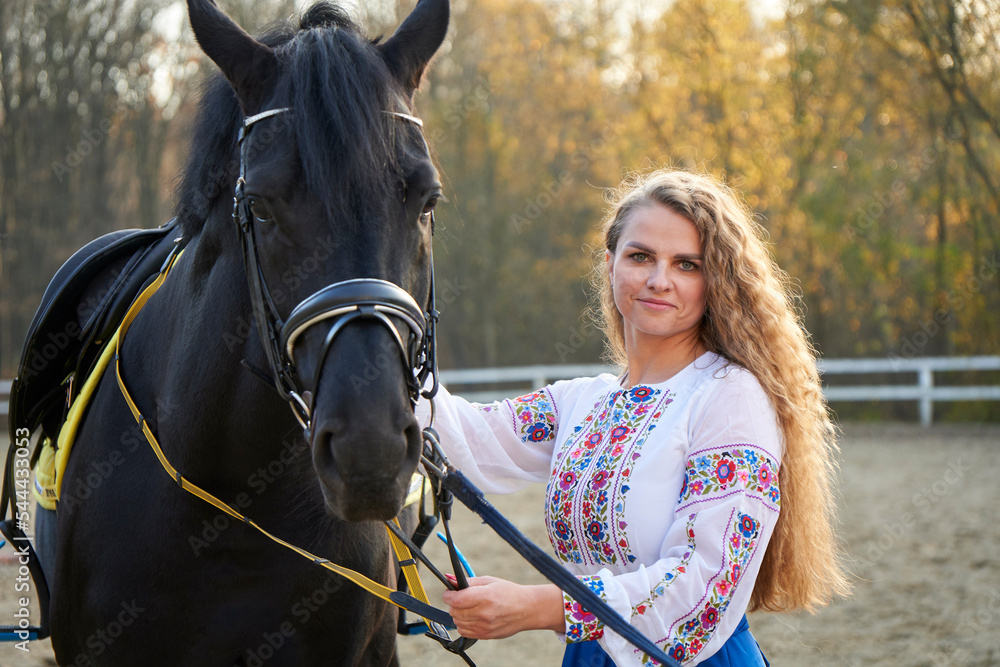 Obraz premium Beautiful young horse woman in a red suit near a stallion. Young sexy woman standing near a horse in a red suit. Beautiful professional female jockey standing near horse. Cute female in a red jacket