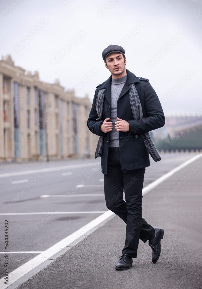 Fashionable handsome male model in black coat and grey flat cap with scarf standing outdoors