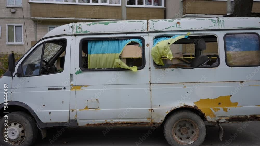 Side view destroyed van on city street at background of bombed residential house. White car in Kyiv Ukraine with national flag flattering in slow motion on side windows Side view destroyed van on city street at background of bombed residential house. White car in Kyiv Ukraine with national flag flattering in slow motion on side windows
