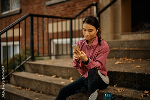 Asian athletic woman text messaging on mobile phone outdoors.