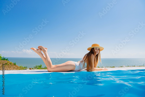 a slender woman in a white swimsuit in a straw hat lies on the pool.