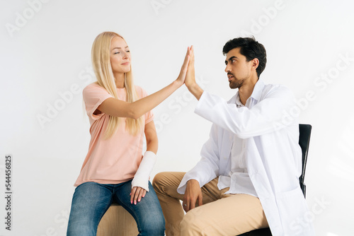 Serious male doctor give high five to pretty blonde female patient with broken hand wrapped in plaster bandage at checkup meeting on white isolated background. Concept of insurance and healthcare.