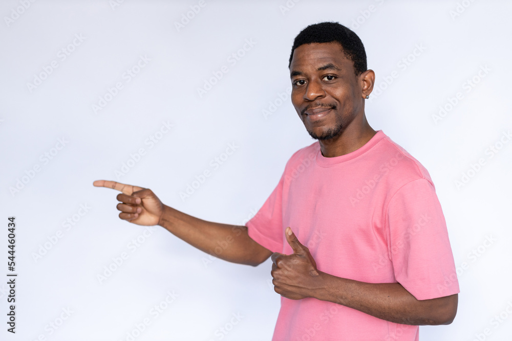 Portrait of pleased African American man pointing finger at ad. Happy young male model with short dark hair in pink T-shirt looking at camera, smiling with thumb up. Advertisement, approval concept Portrait of pleased African American man pointing finger at ad. Happy young male model with short dark hair in pink T-shirt looking at camera, smiling with thumb up. Advertisement, approval concept