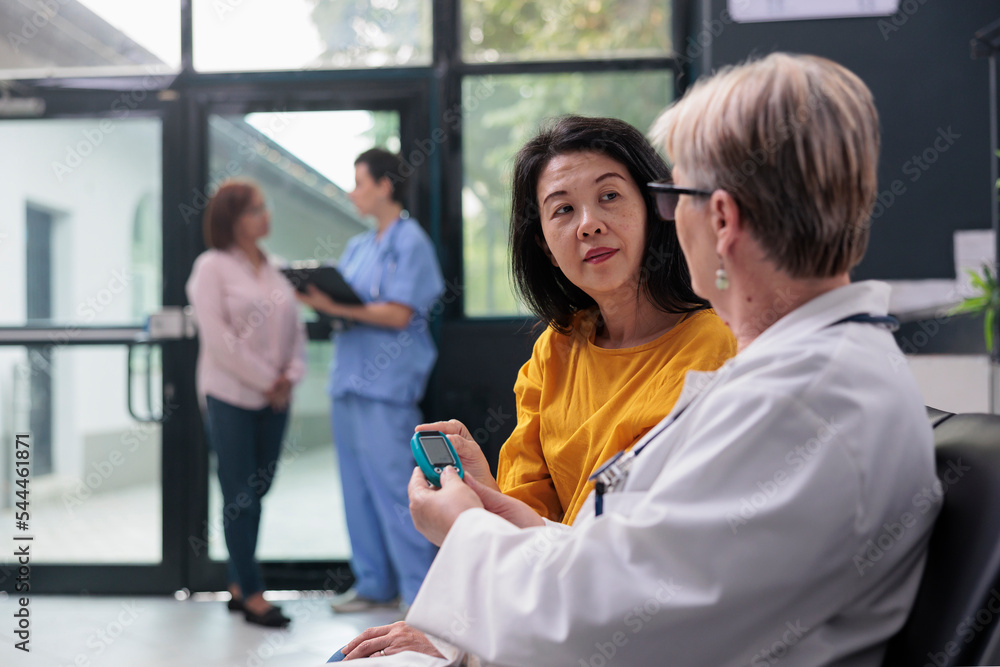 Fototapeta premium Senior medic consulting asian patient with glucometer instrument, measuring insulin and glucose level from blood sample. Doctor and woman doing diabetic control at checkup visit appointment.