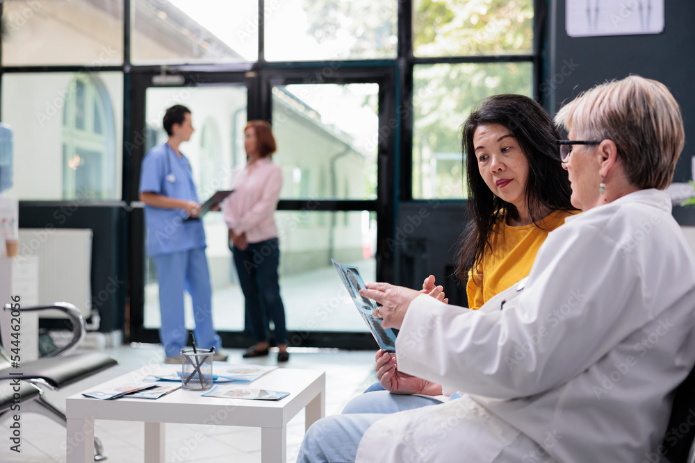 Fototapeta premium Asian patient looking at medical radiography with senior doctor discussing illness diagnosis during consultation in hospital waiting area. Women talking about healthcare service support