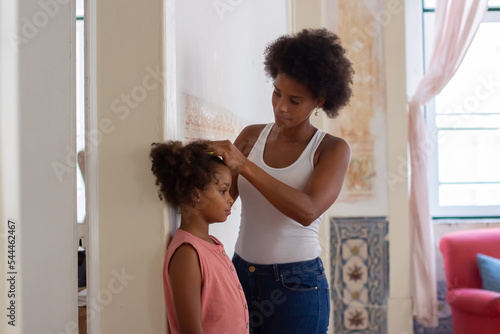 African American woman measuring daughters height. Girl in pink overall standing near wall, mother marking. Childhood, time together concept