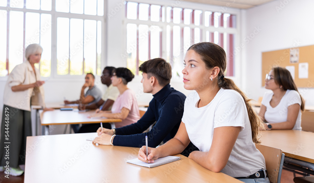 Fototapeta premium Portrait of young girl attentively listening to lecture and taking notes in classroom full of students