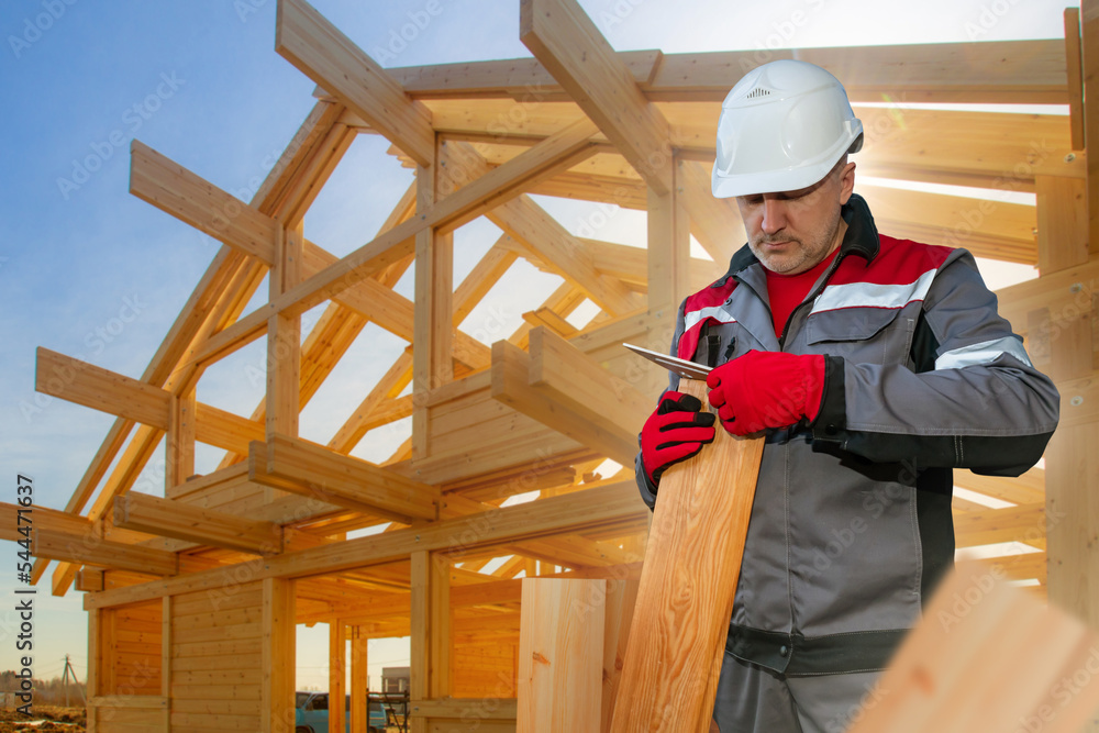 Builder worker. Man with ruler measures board. Worker builds house out of wood. Guy in form of builder. Builder man near frame of wooden house. Concept of checking building materials for quality Builder worker. Man with ruler measures board. Worker builds house out of wood. Guy in form of builder. Builder man near frame of wooden house. Concept of checking building materials for quality