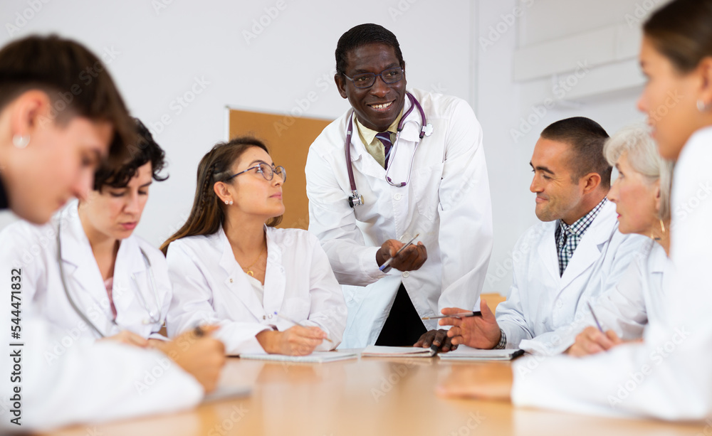 Group of medical workers in white coats sitting at same desk during professional training course, listening to experienced african american lecturer..