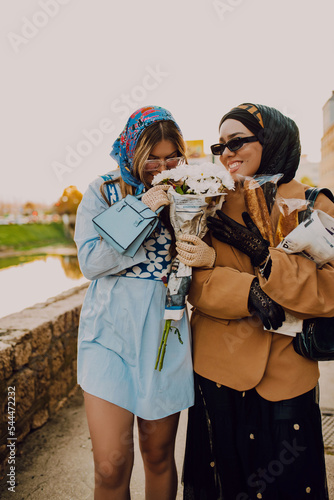 A modern European woman and a Muslim woman with a hijab walking the streets of the city dressed in clothes from the 19th century while carrying newspapers, flowers and bread in their hands.