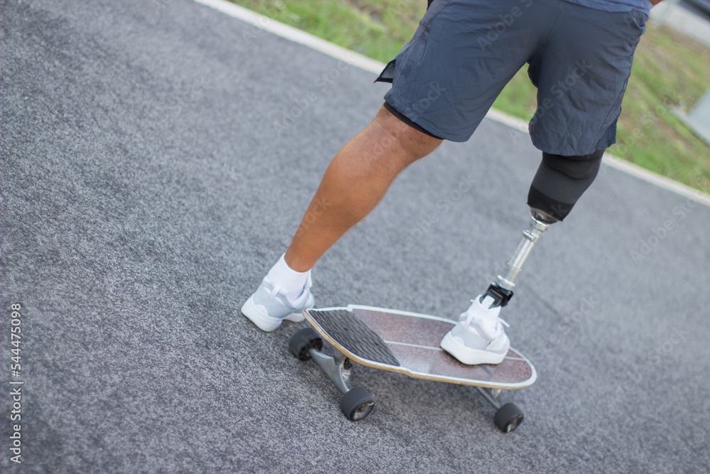 Close-up of sportsman with disability in park on sunny day. Man in casual clothes and sportive protection riding skateboard. Sport, disability, training concept Close-up of sportsman with disability in park on sunny day. Man in casual clothes and sportive protection riding skateboard. Sport, disability, training concept