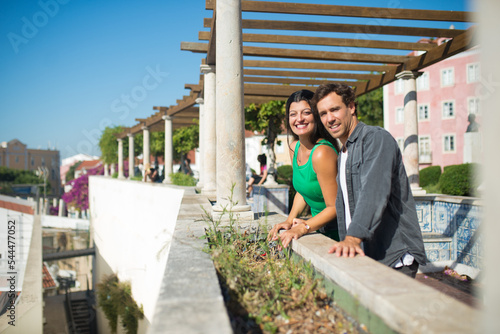 Portrait of smiling man and woman travelling around city. Happy man and woman standing on summer alley with columns looking at city houses and views from height. Relations, love and travelling concept