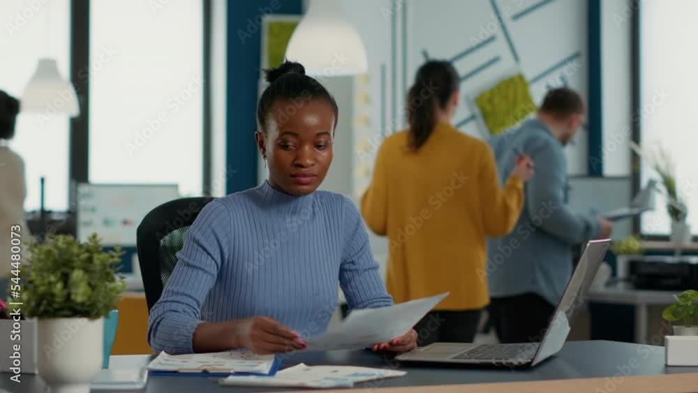 African american startup employee holding business papers with charts looking at laptop screen comparing data. Woman in busy office analyzing marketing statistics and annual turnover. African american startup employee holding business papers with charts looking at laptop screen comparing data. Woman in busy office analyzing marketing statistics and annual turnover.