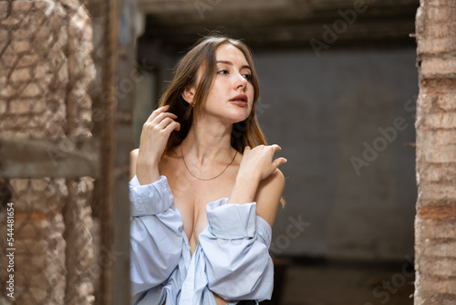 Portrait of young desirable woman taking off her shirt, demonstrating naked shoulders, covering her breast in abandoned building.