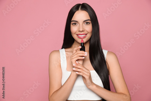 Young woman with beautiful makeup holding glossy lipstick on pink background