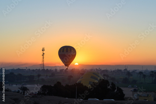 Colorful hot air balloons fly over the green field at sunrise in Valley of the Kings and red cliffs western bank of the Nile river- Luxor- Egypt