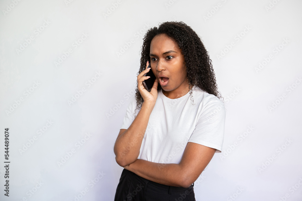 Portrait of astonished young woman talking on mobile phone. African American woman wearing white T-shirt listening to friend with open mouth. Mobile communication and surprise concept Portrait of astonished young woman talking on mobile phone. African American woman wearing white T-shirt listening to friend with open mouth. Mobile communication and surprise concept