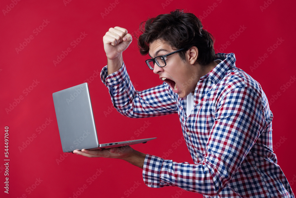 Furious angry young man shouting at his laptop, red studio background Furious angry young man shouting at his laptop, red studio background