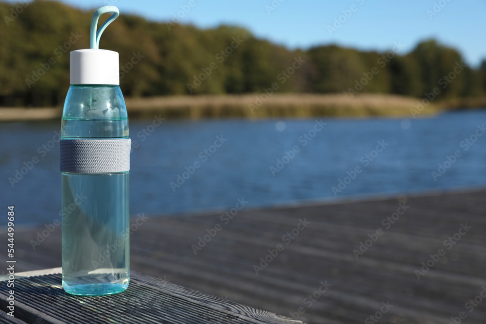 Glass bottle with water on wooden pier near river outdoors. Space for text
