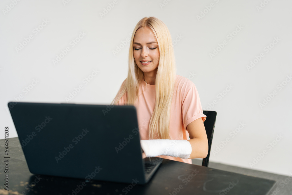 Studio portrait of cheerful business woman with broken arm wrapped in white plaster bandage working typing on laptop computer, looking to screen, sitting at desk on light background. Studio portrait of cheerful business woman with broken arm wrapped in white plaster bandage working typing on laptop computer, looking to screen, sitting at desk on light background.