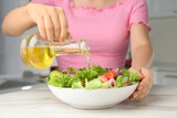 Woman pouring oil from jug into bowl with salad in kitchen, closeup
