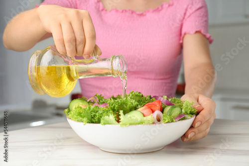 Woman pouring oil from jug into bowl with salad in kitchen, closeup