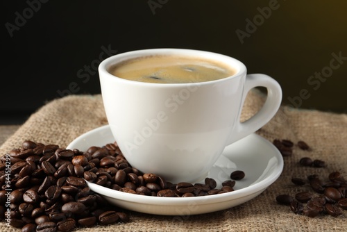 Cup of hot aromatic coffee and roasted beans on sackcloth against dark background