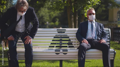 Mature men in formal suit and safety mask relaxing on bench inpark keeping social distance. Realtime photo